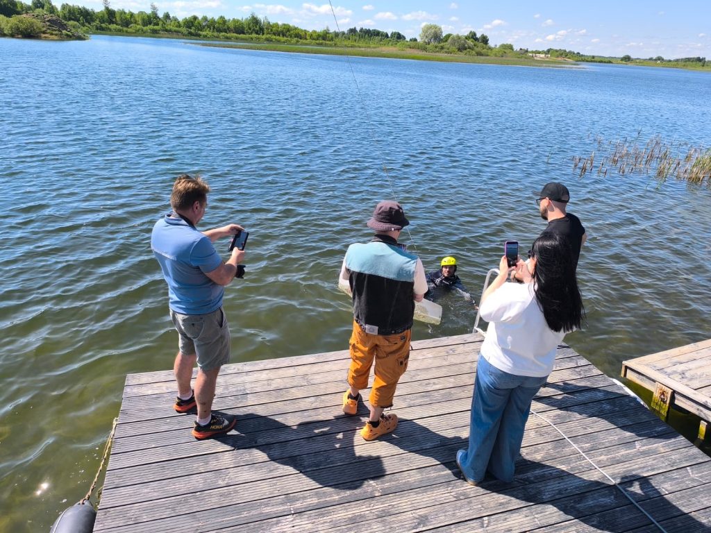 Group of people on a wooden dock by a lake, watching a person in water wearing a helmet. Some individuals are taking photos with their phones, while others are engaged in conversation. Lush greenery and blue sky are visible in the background, indicating a sunny day. Group of people on a wooden dock by a lake, watching a person in water wearing a helmet. Some individuals are taking photos with their phones, while others are engaged in conversation. Lush greenery and blue sky are visible in the background, indicating a sunny day.