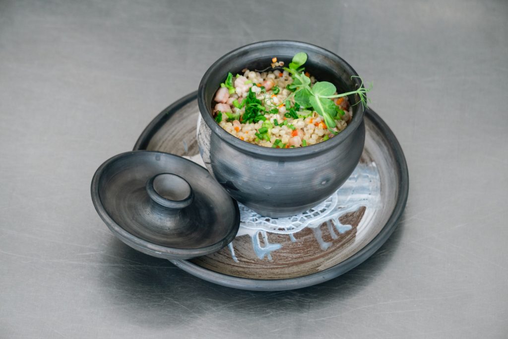 A traditional black clay pot filled with colorful vegetable and grain dish, garnished with fresh herbs, served on a decorative plate.