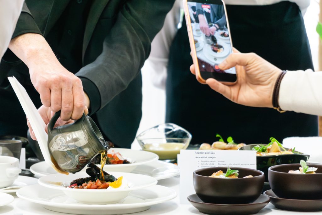 A server pours sauce from a black jug onto a gourmet dish while a diner captures the moment with a smartphone. In the background, elegantly presented plates and a menu card are visible, showcasing fine dining and culinary artistry.