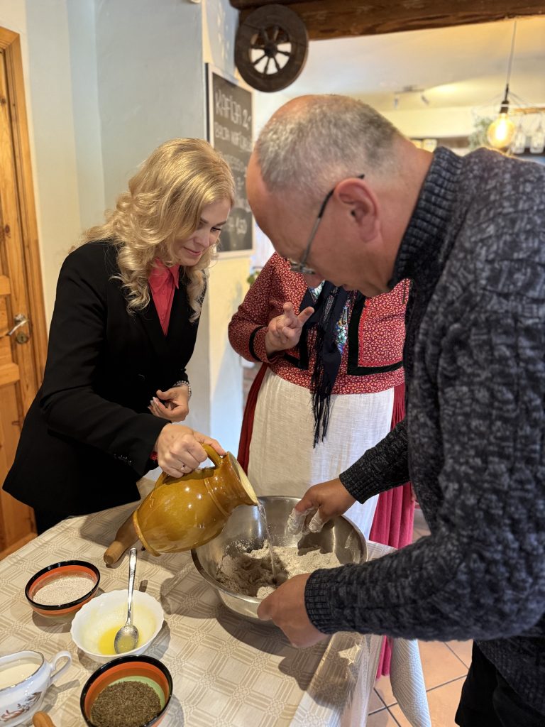 Group of people engaged in cooking activity, with a woman pouring liquid into a mixing bowl while a man stirs flour and other ingredients. The setting features a rustic kitchen with wooden accents and various bowls of ingredients on the table.