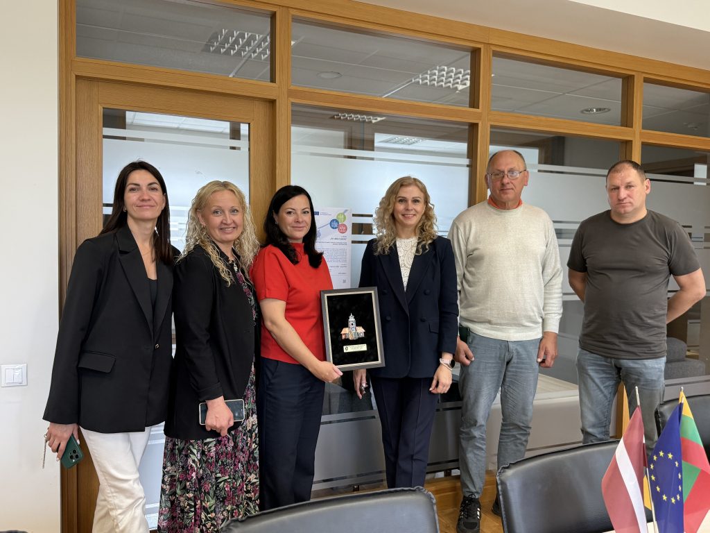 Group of professionals standing together in an office setting, showcasing a framed award. The team includes three women and three men, with flags of Latvia, the European Union, and Bulgaria visible on the table. The image highlights collaboration and achievement in a business environment.