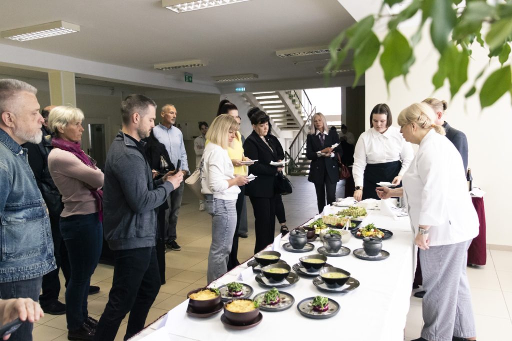 Group of people attending a culinary event, observing a chef presenting a variety of dishes on a table, including soups and salads, in a modern indoor setting.
