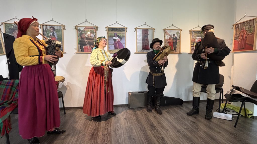 Group of four musicians in traditional attire performing with instruments, including a bagpipe and drums, in front of framed photographs depicting cultural heritage. The setting highlights folk music and traditions.