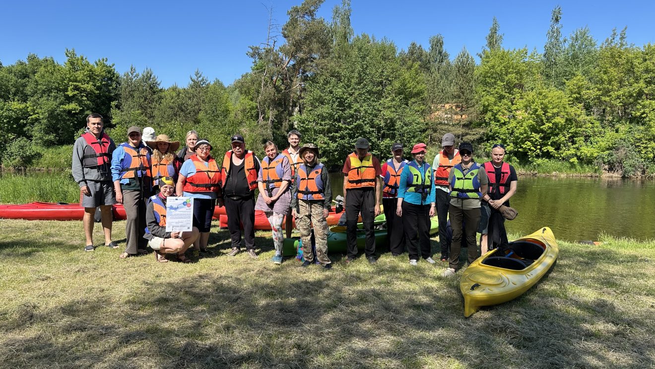 Group of 20 people wearing life jackets gathered by a river with kayaks in the background, participating in a kayaking event on a sunny day.