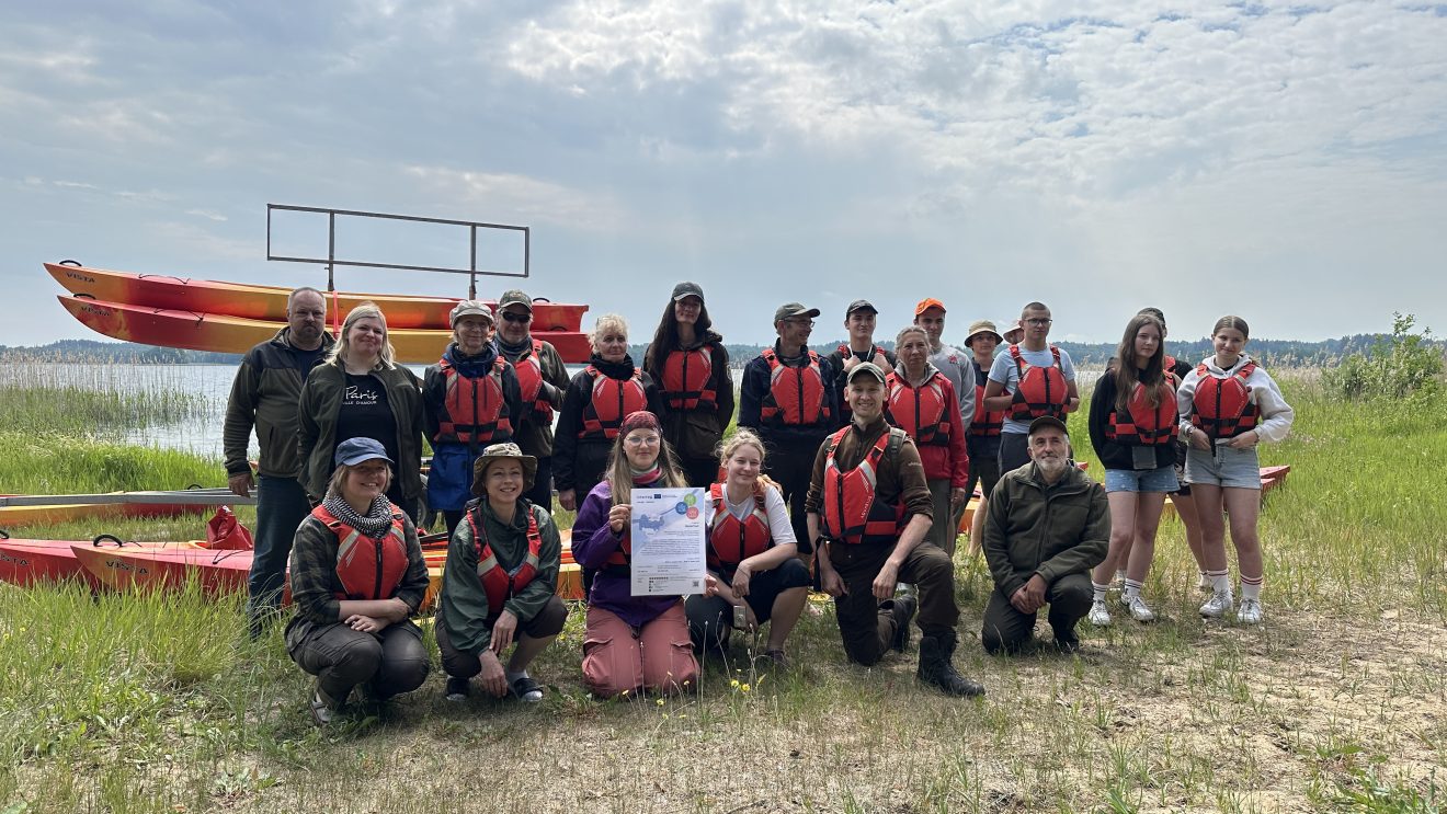 Group photo of participants in a kayaking event, wearing life jackets, with kayaks in the background. The setting is a grassy area near a lake, showcasing a diverse group celebrating their achievements.