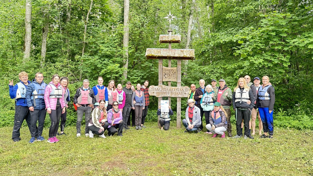 Group of hikers in life jackets gathered around a wooden sign marking the northernmost point of Lithuania, surrounded by lush greenery and trees.