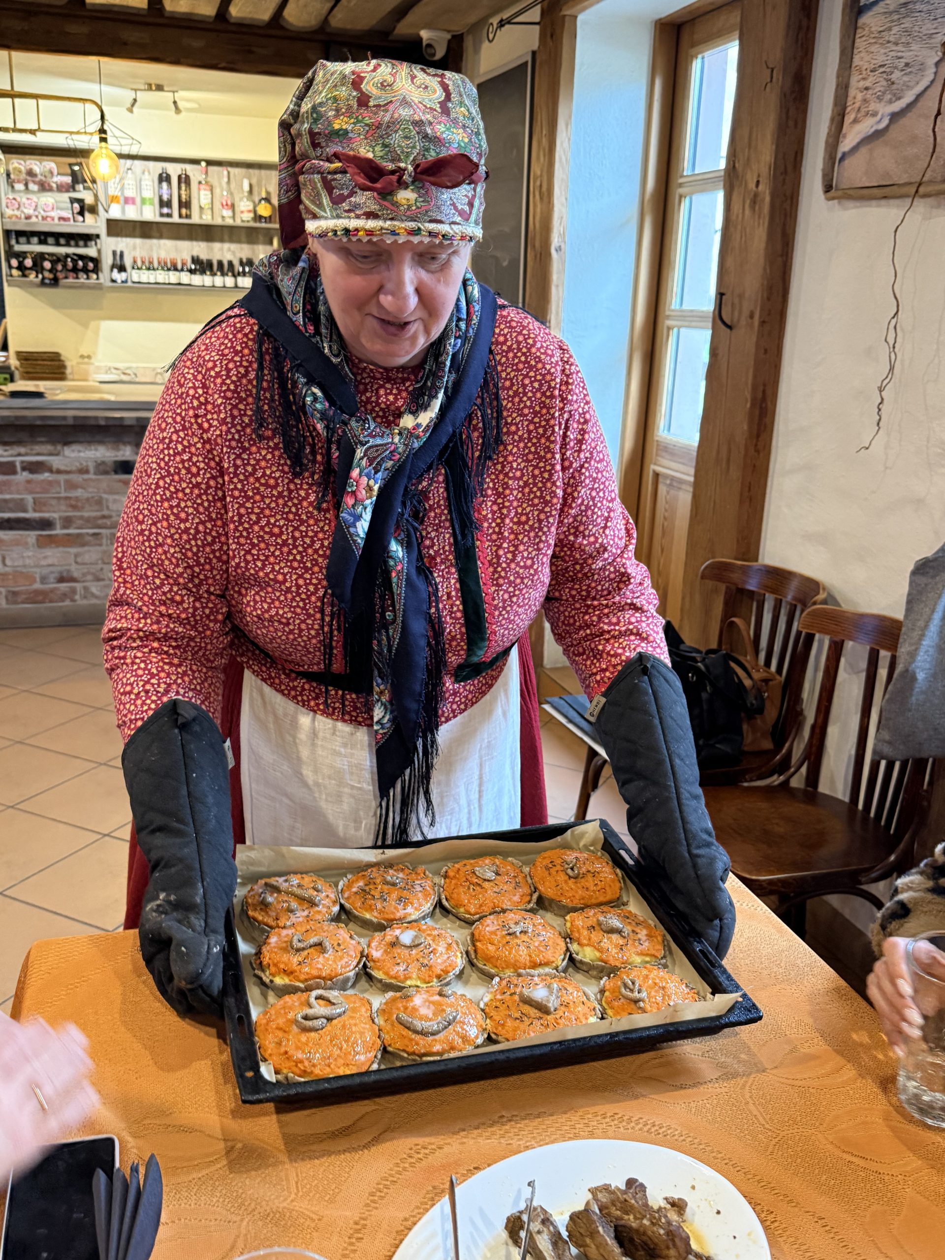 A woman in traditional attire serves a tray of freshly baked pastries topped with orange glaze and garnished with herbs, in a cozy dining setting. The background features a rustic bar area with bottles and a warm, inviting atmosphere.
