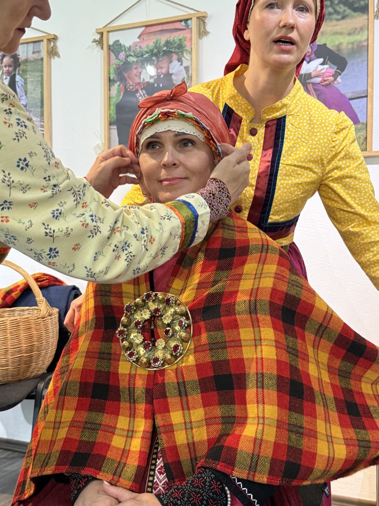 Two women are engaged in a traditional costume fitting, showcasing vibrant cultural attire. One woman, wearing a red and yellow plaid shawl adorned with a decorative brooch, is having her headscarf adjusted by another woman in a floral blouse. The background features framed photos of individuals in similar traditional dress, highlighting the cultural significance of the attire.