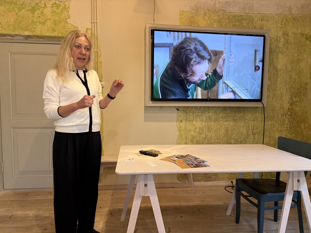 A woman with long blonde hair gestures while presenting in a room with green accent walls. A television displays a close-up of a person working on a wooden frame. A table in the foreground holds brochures and a remote control, contributing to a professional and engaging atmosphere.