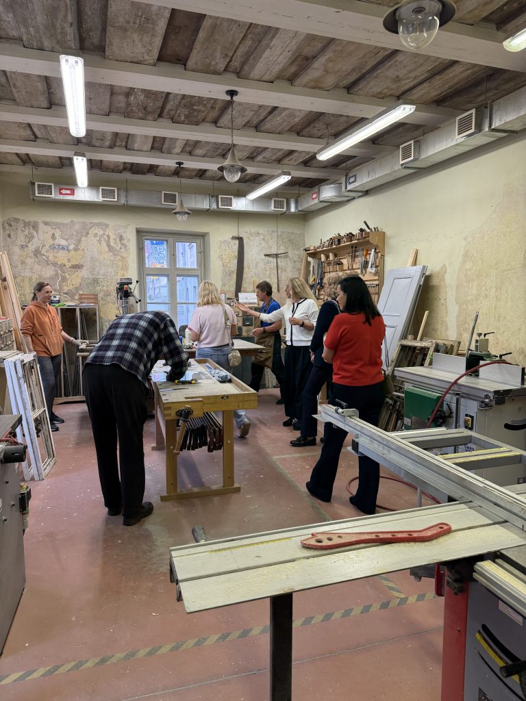 Group of people participating in a woodworking workshop, surrounded by tools and materials in a well-lit studio. The space features wooden beams and various woodworking equipment, with individuals engaged in hands-on learning and collaboration.