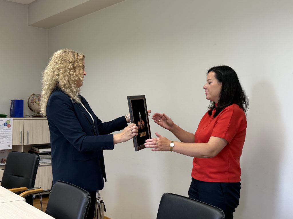 Two women are engaged in a professional exchange, with one presenting a framed item to the other in a modern office setting. The woman in a black blazer has curly hair, while the woman in a red shirt appears to be receiving the frame. A globe and office materials are visible in the background, indicating a collaborative environment.