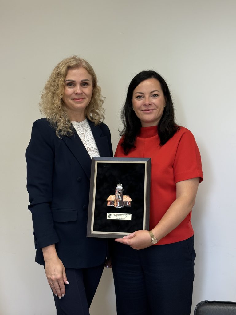 Two women stand side by side, one in a navy suit and the other in a red top, both smiling while holding a framed award featuring a model of a building. The background is plain and neutral-colored, emphasizing their expressions and the award.