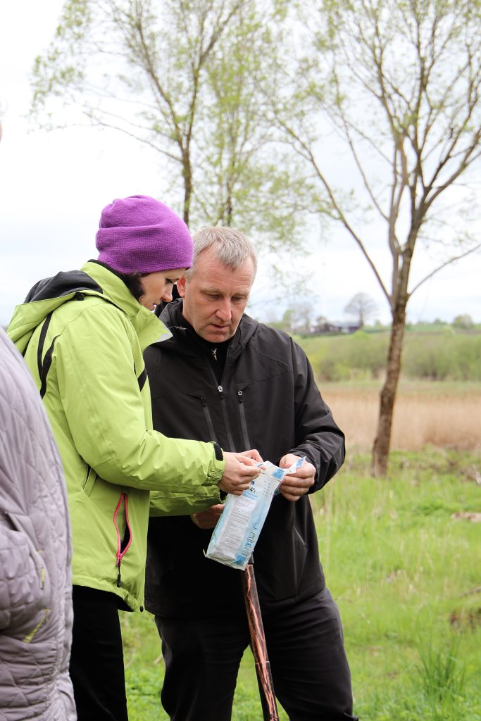 Two adults engaged in a discussion outdoors, one wearing a green jacket and purple beanie, while the other is in a black jacket. They are examining a package together, surrounded by trees and greenery, indicating a nature-related activity or workshop.