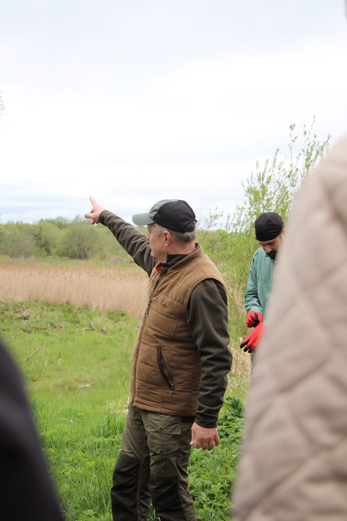 A man in a brown vest gestures while leading a nature walk, with lush greenery and tall grass in the background. Another participant in a green jacket is seen adjusting gloves, indicating an outdoor educational activity focused on environmental awareness.