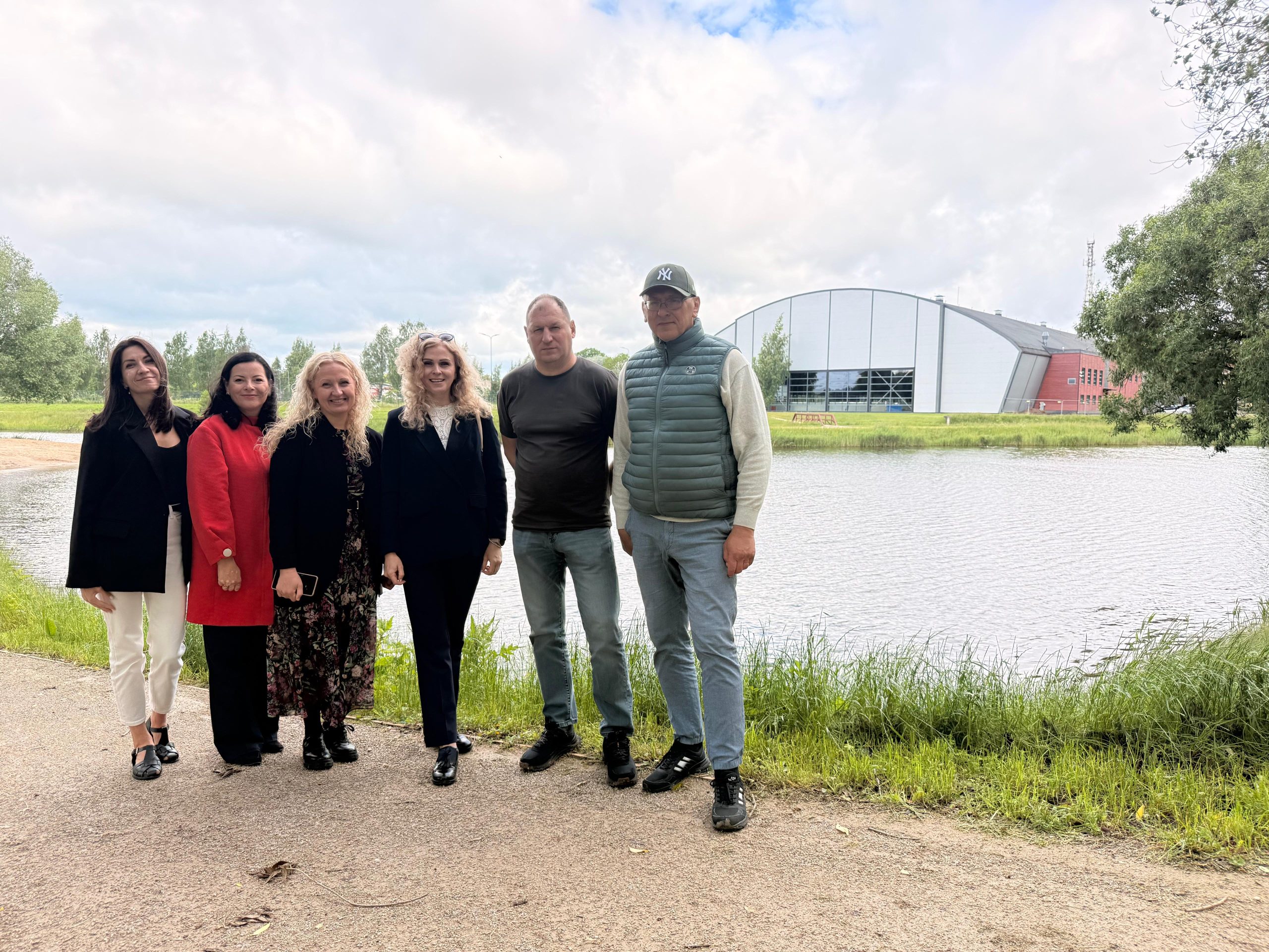 Group of six professionals posing outdoors by a lake, with a modern building in the background. The individuals are dressed in business attire, showcasing a mix of styles and colors, while the surrounding greenery and cloudy sky create a serene atmosphere.