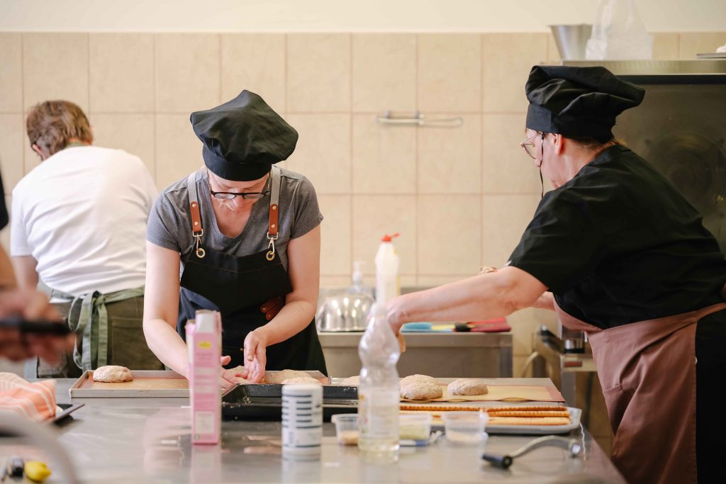 Bakers in a kitchen preparing dough, wearing black chef hats and aprons, with various baking ingredients and tools on the counter.