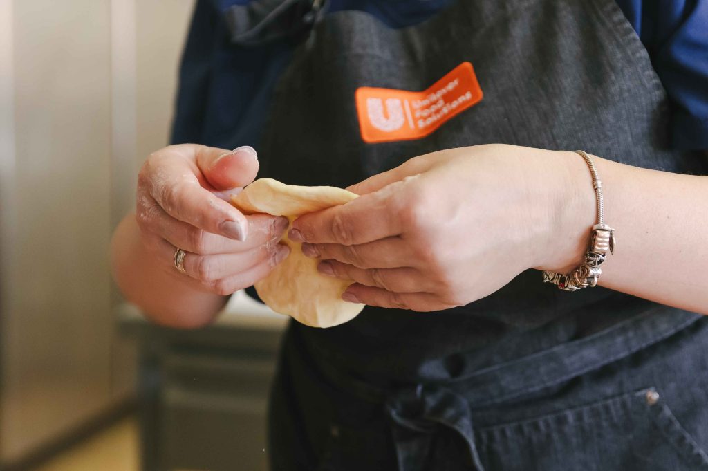 Hands of a person in a dark apron working with dough, shaping it by hand in a kitchen setting, highlighting culinary skills and food preparation techniques.