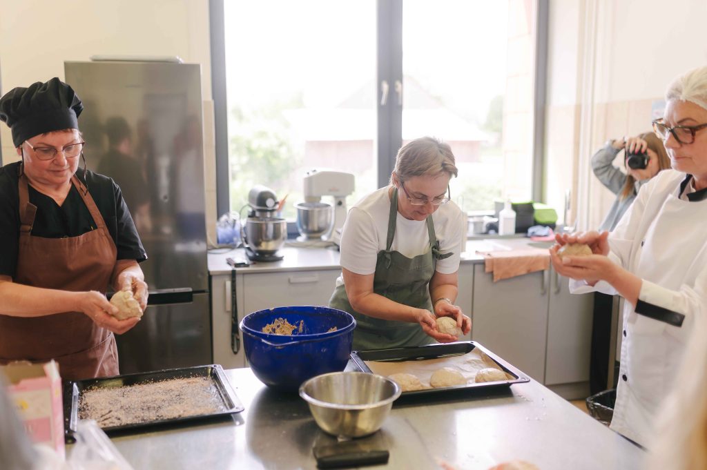 Three women in a modern kitchen are preparing dough for baking. One woman is shaping dough balls, while another holds a formed piece of dough. A blue mixing bowl with dough and baking trays are visible on the counter, showcasing an active baking environment.