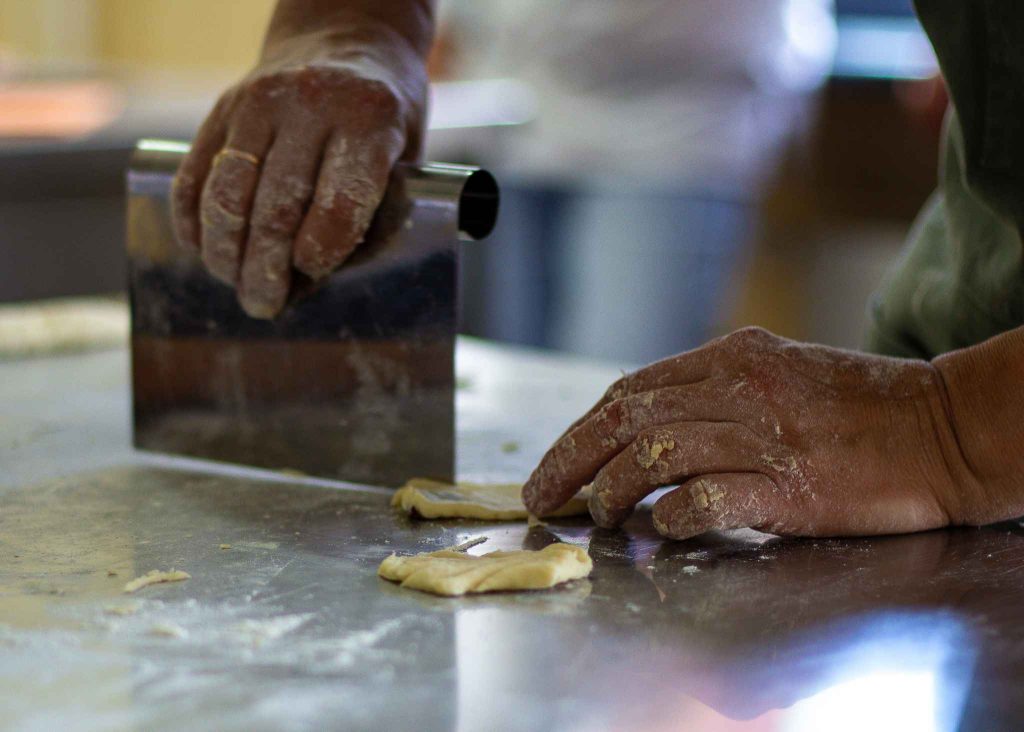 Hands covered in flour using a metal bench scraper to cut dough on a stainless steel surface, showcasing the art of baking and pastry preparation.