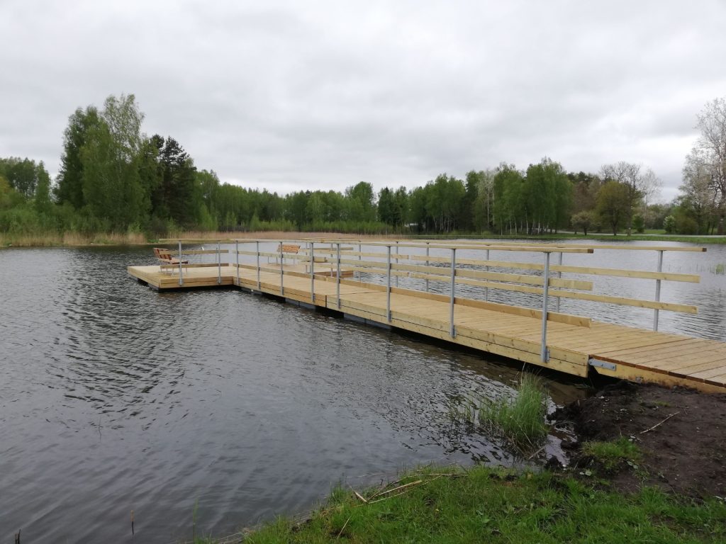 Wooden dock extending into a calm lake surrounded by lush greenery and trees under a cloudy sky, featuring benches for relaxation.