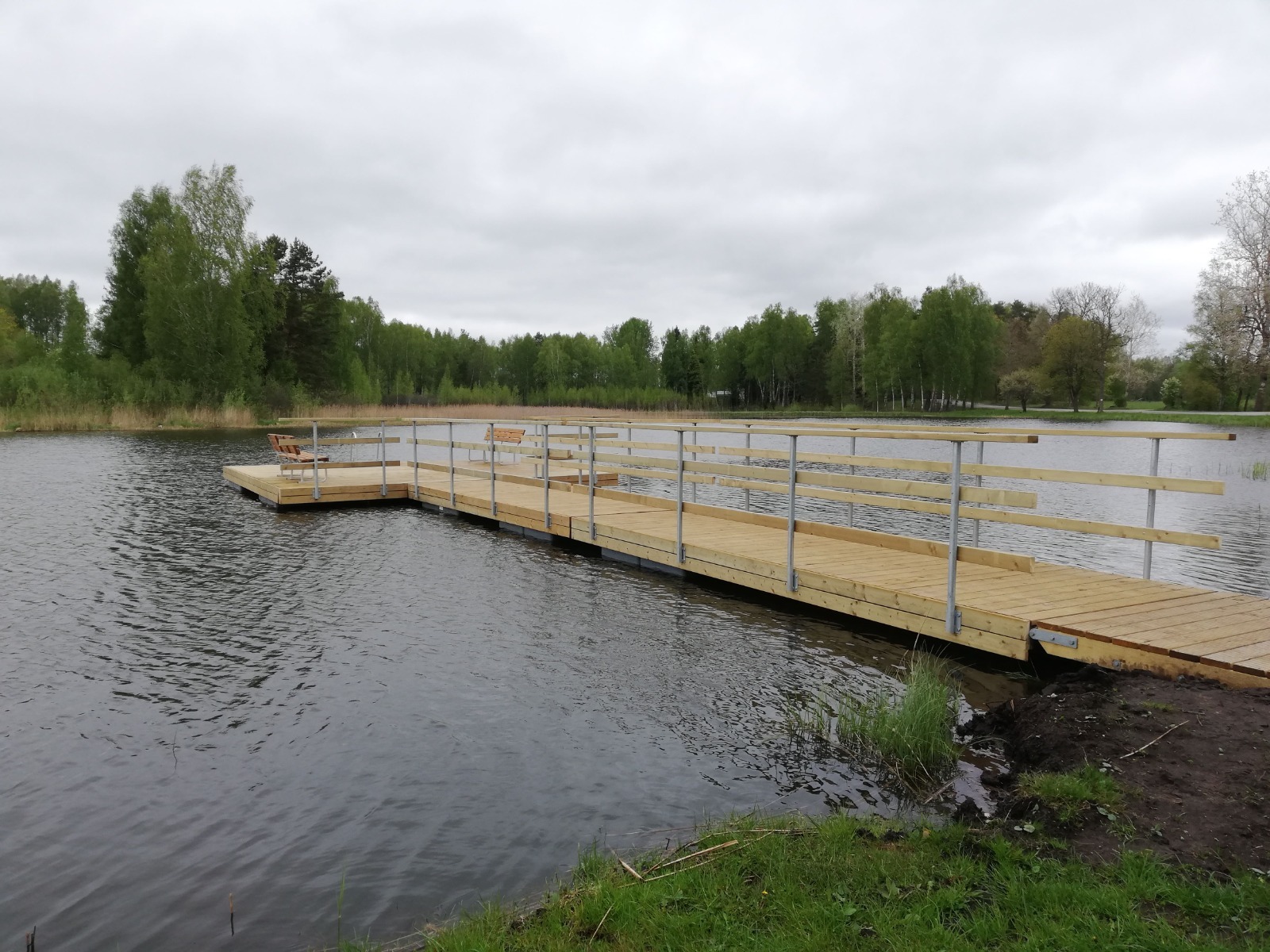 Wooden dock extending into a calm lake surrounded by lush greenery and trees under a cloudy sky, featuring benches for relaxation.
