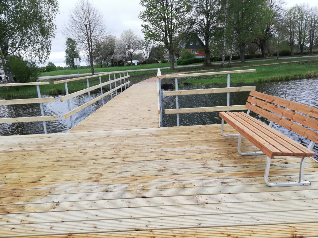Wooden dock extending over a calm body of water, featuring a metal railing and a wooden bench for seating, surrounded by greenery and trees. Ideal location for relaxation and enjoying nature.