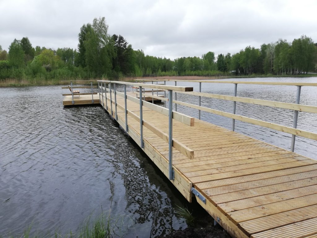 Wooden dock extending over a calm lake, surrounded by lush greenery and cloudy skies, ideal for fishing and nature activities.