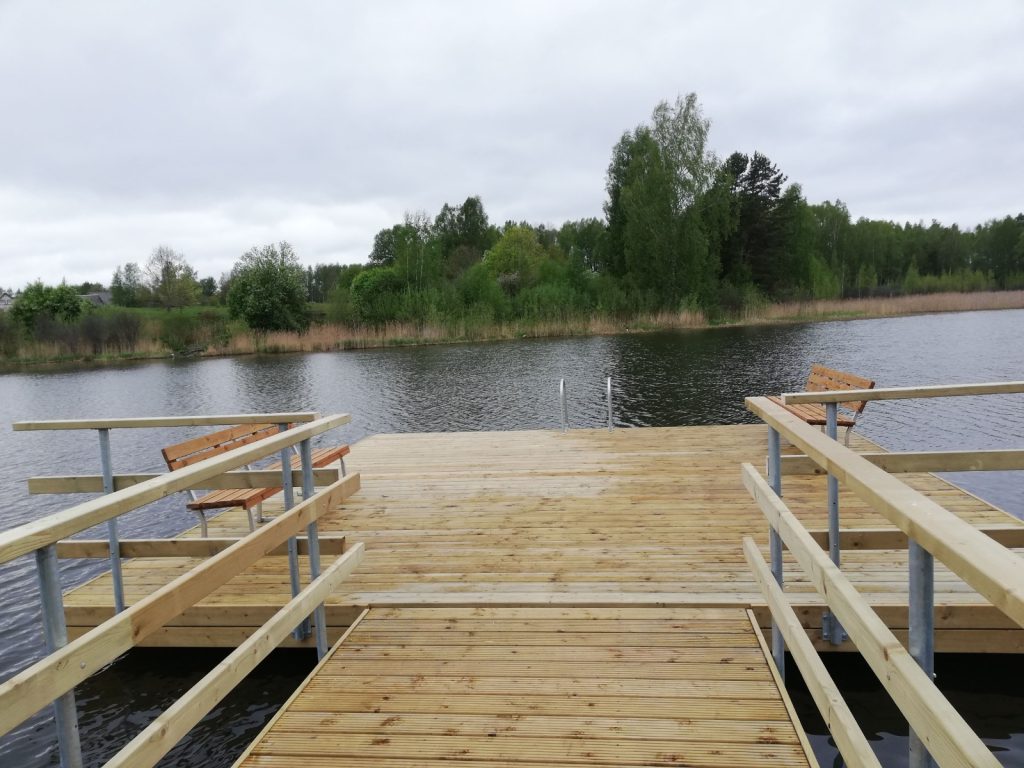 A wooden dock extending over a calm lake, featuring two benches and surrounded by lush greenery and reeds under a cloudy sky.