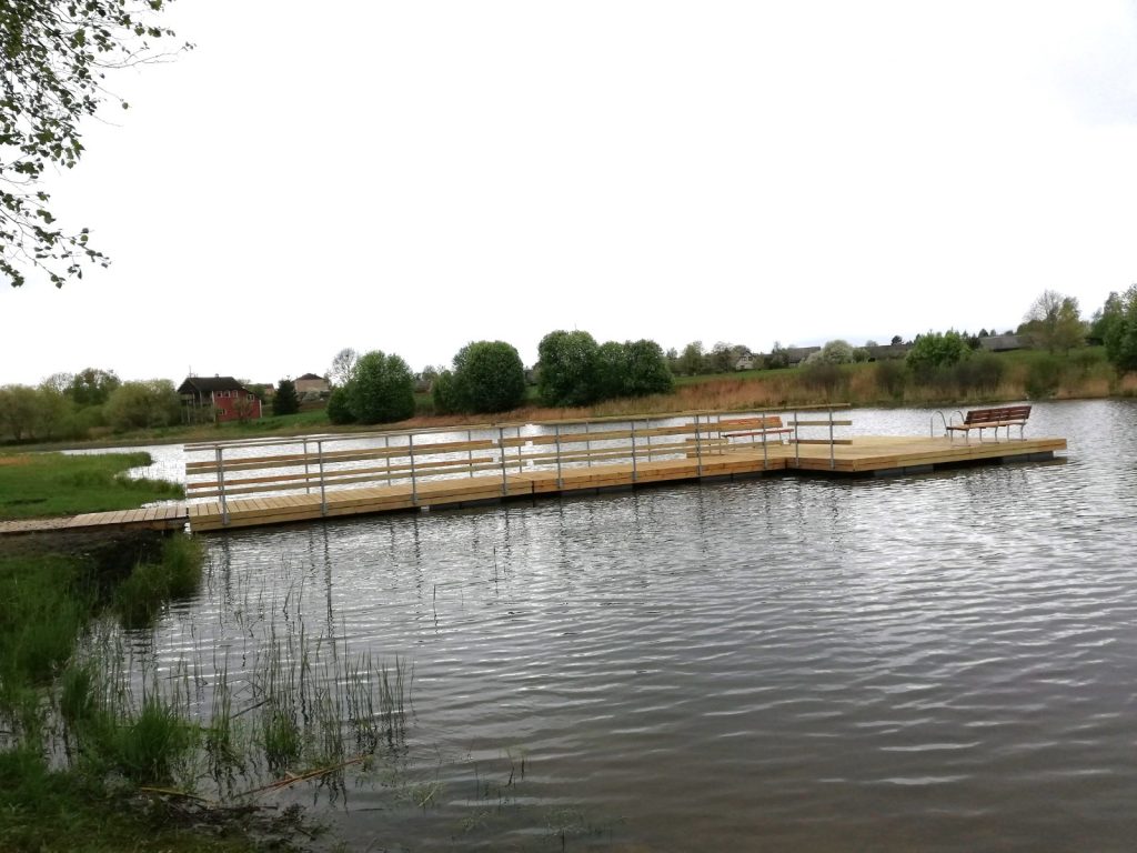 Wooden dock extending over a tranquil lake, surrounded by greenery and benches for relaxation, with a cloudy sky in the background. Ideal for nature walks and outdoor leisure activities.