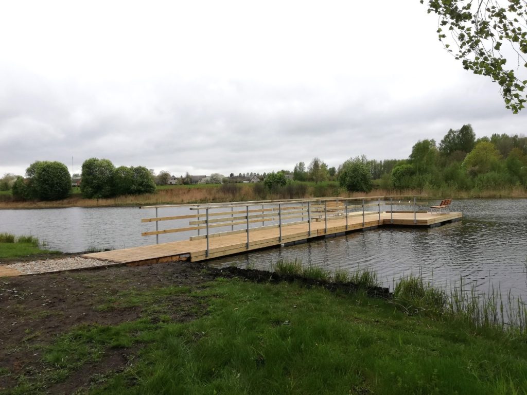 Wooden dock extending into a calm lake surrounded by greenery, with cloudy skies overhead. Ideal for fishing or enjoying nature.
