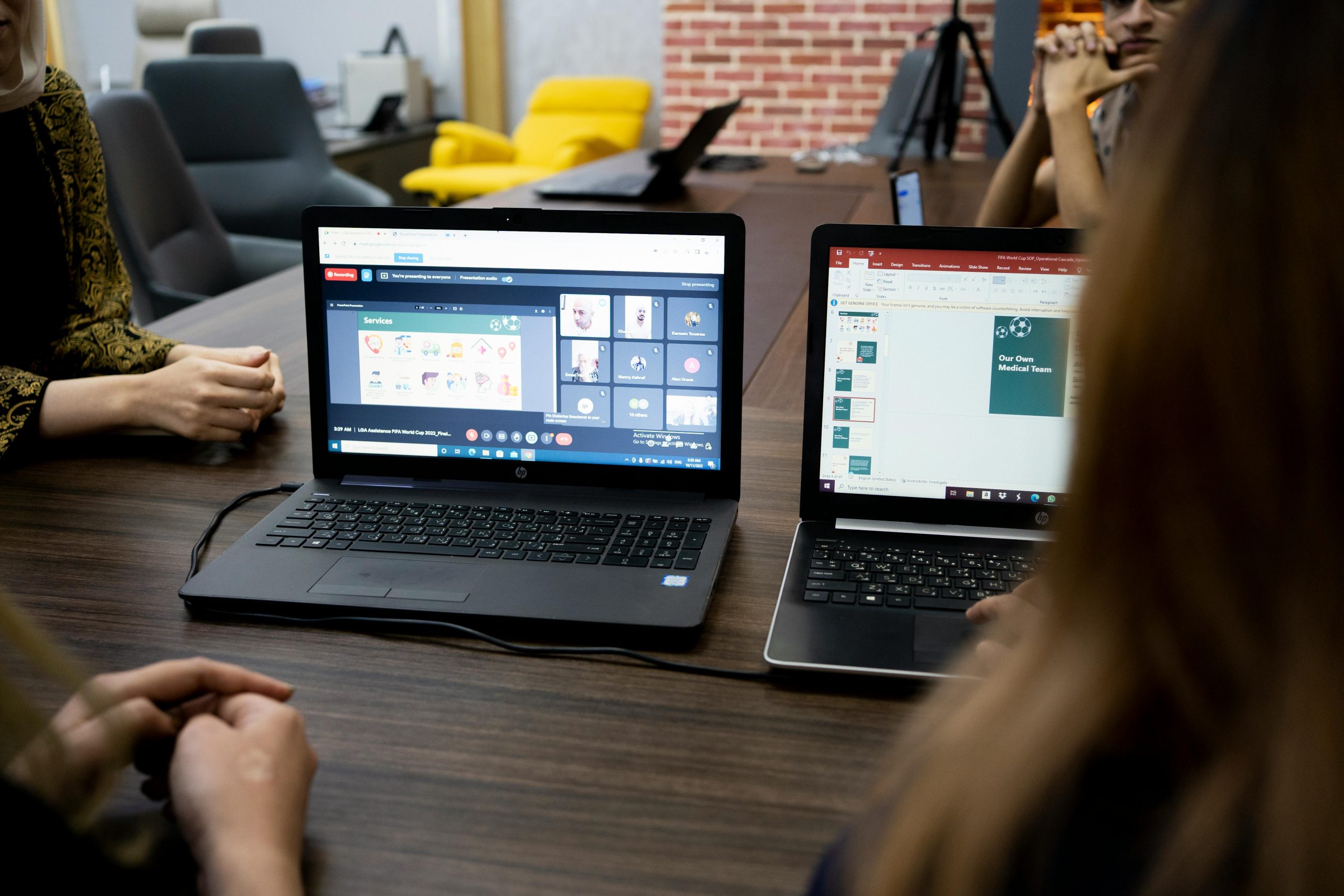 Two laptops are open on a conference table, showing a virtual meeting on one screen and a presentation on the other. Participants are engaged in a collaborative work environment. The setting suggests a professional atmosphere focused on teamwork and digital communication.