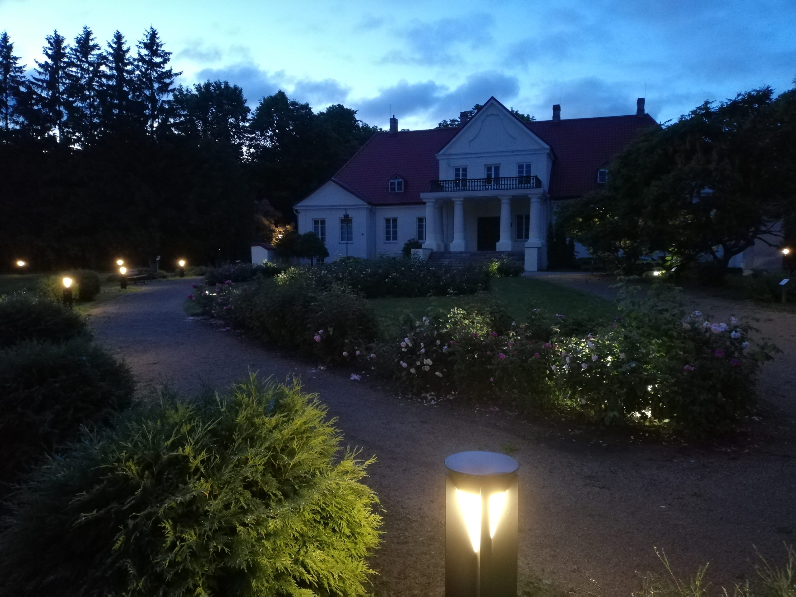 Footpath lighting illuminating the pathway in Joniškėlis Manor Park at dusk, showcasing landscaped gardens and the manor house in the background.