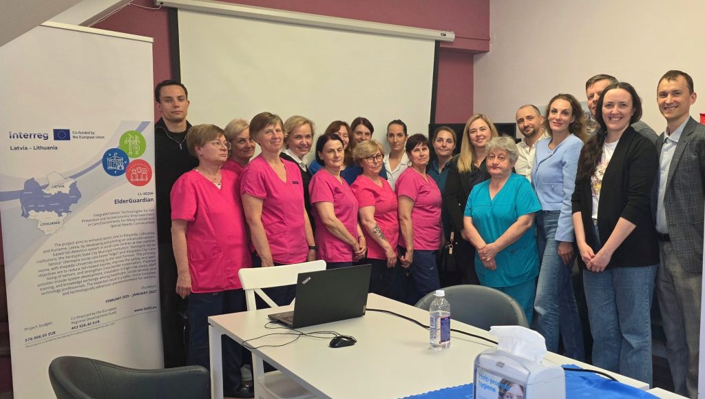 Group of healthcare professionals in pink and blue uniforms gathered for the initial training of the "ElderGuardian" project, focused on fall prevention and support for the elderly in care environments. A laptop and water bottle are visible on the table, with a project banner in the background detailing the initiative's goals and funding sources.