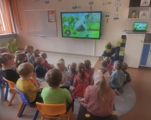 Children watching an educational video in a classroom, seated on colorful chairs, with a large screen displaying animated content. The room is bright and organized, featuring playful decor and soft seating areas for a learning environment.