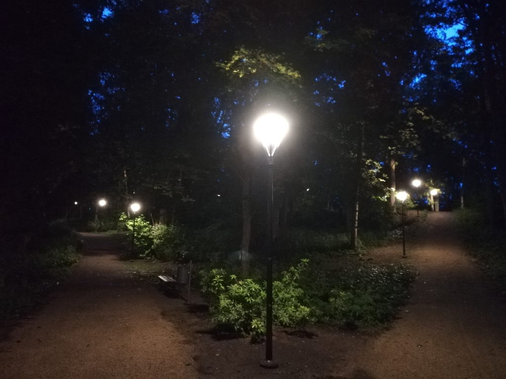 Footpath lighting illuminating pathways in Joniškėlis Manor Park at dusk, surrounded by trees and greenery.
