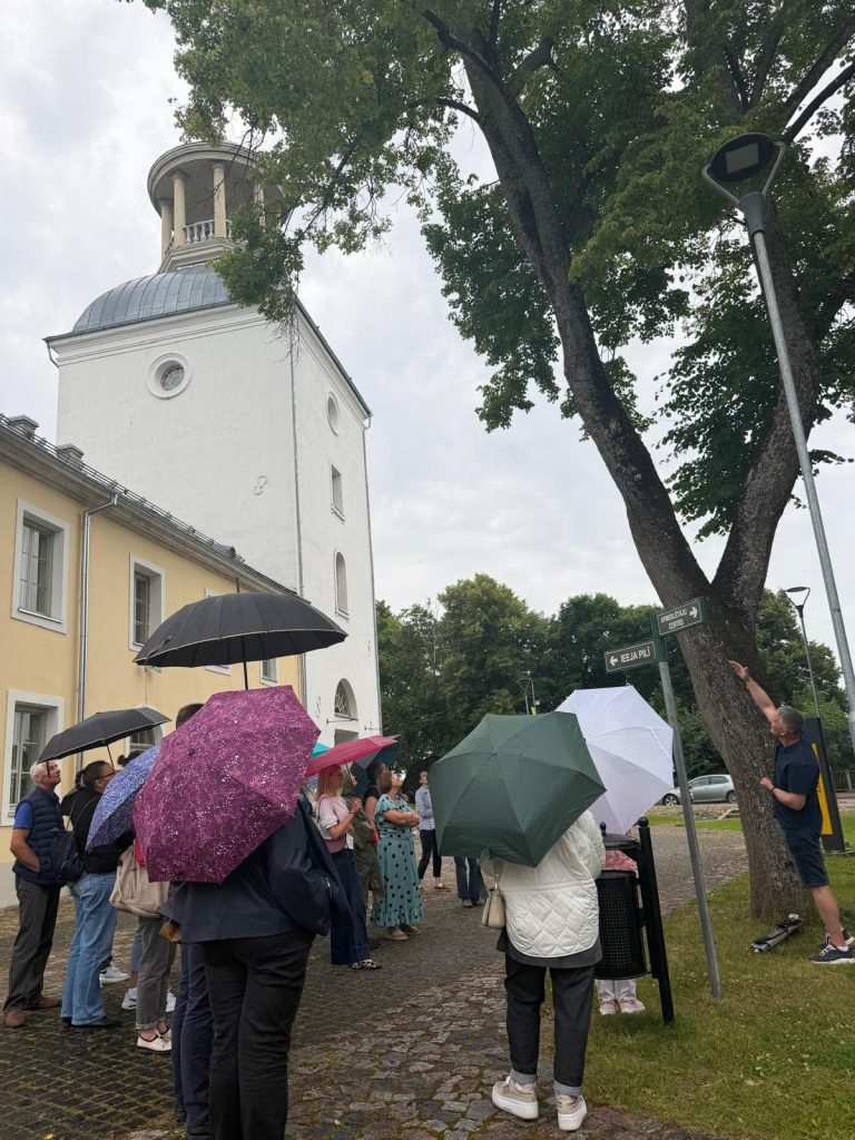 Participants gather under umbrellas outside Jēkabpils History Museum during a lecture and practical workshop on sustainable park management, with a speaker pointing towards a tree.