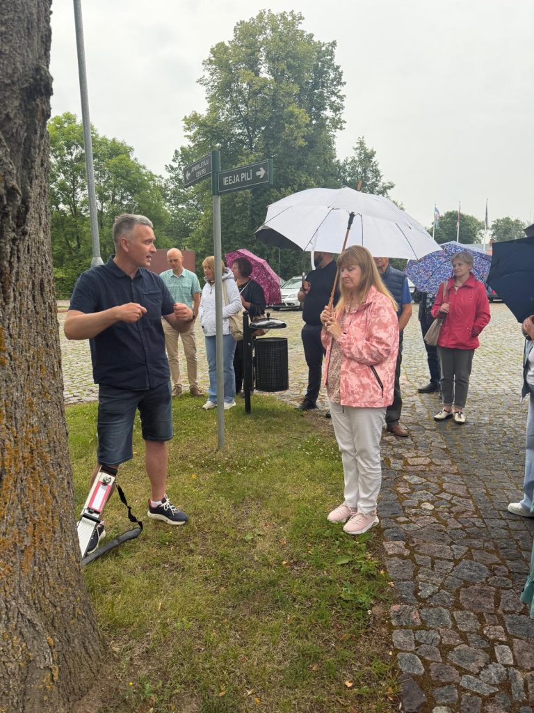 Participants engage in a practical workshop on sustainable park management at Jēkabpils History Museum, with a speaker demonstrating techniques while attendees listen under umbrellas on a rainy day.