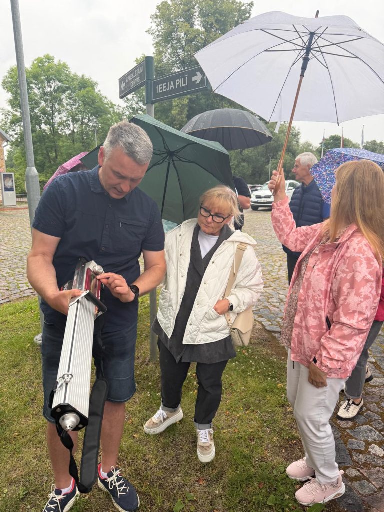 Participants at the Jēkabpils History Museum engage in a practical workshop on sustainable park management, observing a demonstration with equipment while holding umbrellas due to rainy weather.