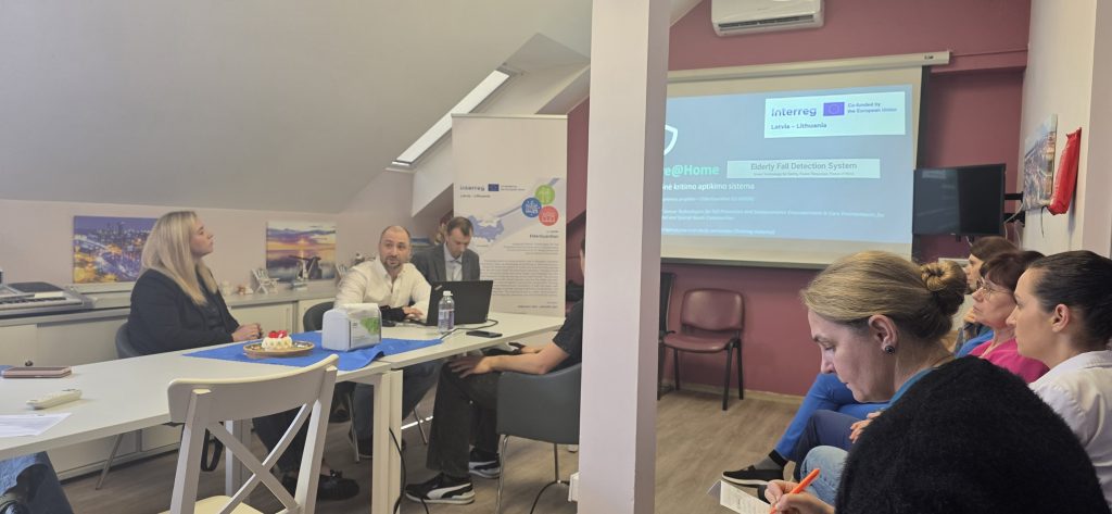 Meeting room scene featuring a training session for the "ElderGuardian" project, with personnel engaged in discussions about the Elderly Fall Detection System. The setting includes a presentation on smart technology for elderly care, with participants taking notes and interacting around a table.