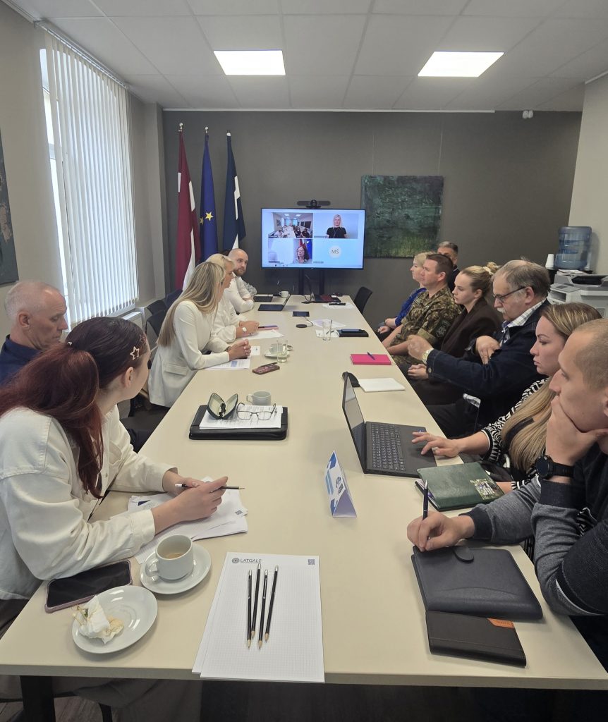 Meeting in progress with participants discussing strategies around a conference table, featuring laptops, notepads, and refreshments. National flags of Latvia and the European Union are displayed, with a video conference screen showing remote attendees.