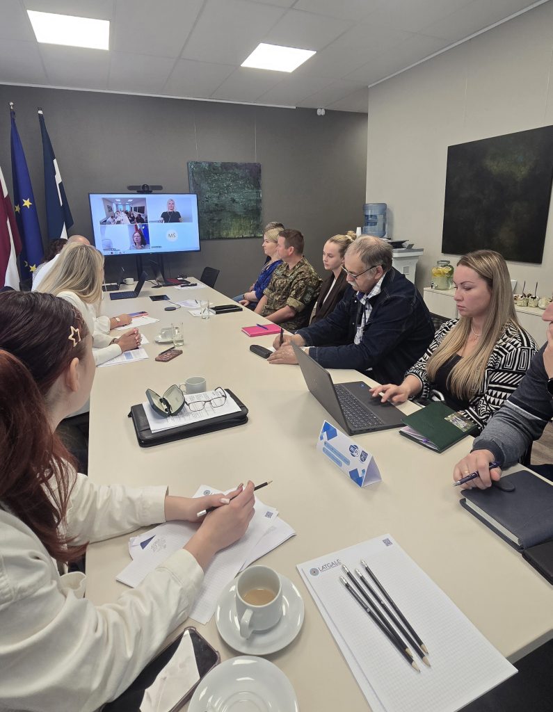 Meeting room with participants engaged in a video conference, discussing various topics. The setting features a modern conference table, laptops, notepads, and refreshments, with flags of Latvia and the European Union displayed in the background.