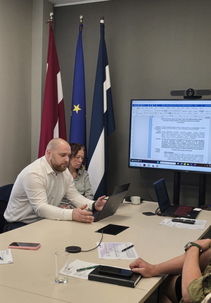 Meeting in a conference room featuring the flags of Latvia, the European Union, and Finland. Two professionals are engaged in a discussion while using laptops, with a large screen displaying a document. The setting includes various documents, a glass of water, and electronic devices on the table.