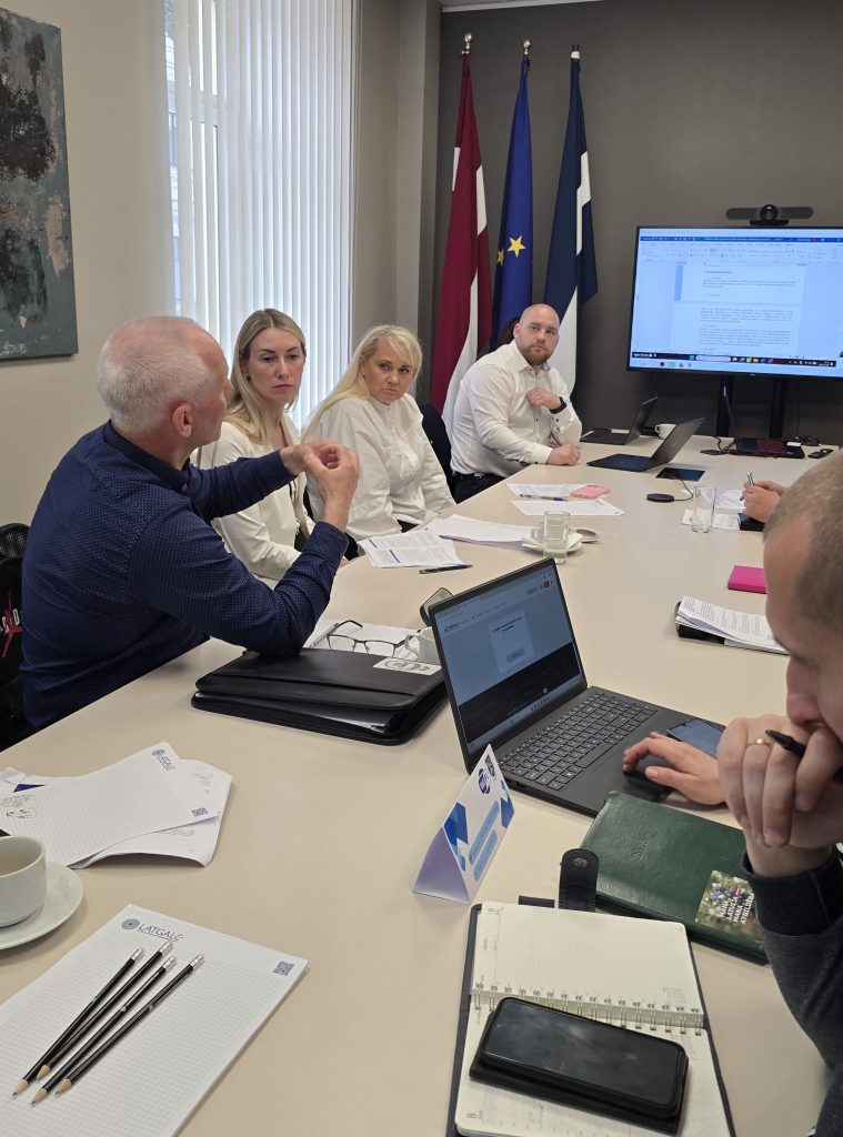 Business meeting in a conference room with participants discussing over documents and laptops, featuring flags of Latvia, the European Union, and Finland in the background.