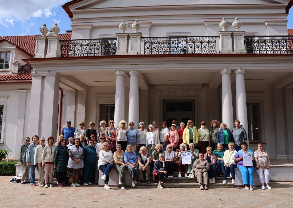 Group photo of a large gathering of adults, predominantly women, standing in front of a historic building with columns and a balcony. The attendees are dressed in casual clothing and smiling, showcasing a sense of community and togetherness. The setting is outdoors with a blue sky and clouds visible above.