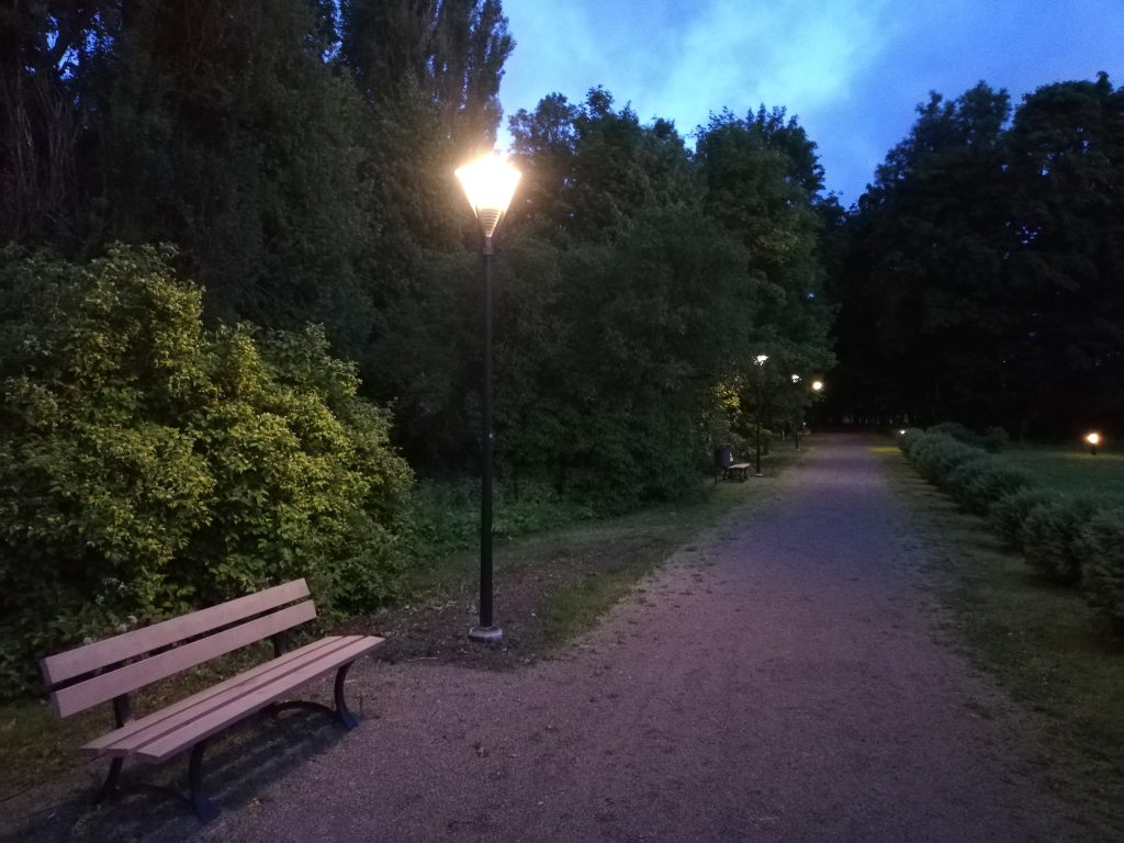 Footpath lighting illuminating the pathway in Joniškėlis Manor Park at dusk, featuring benches and lush greenery along the sides.