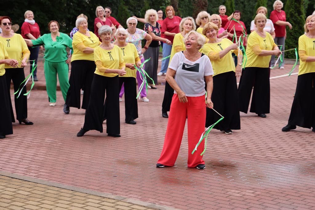 Group of senior women participating in a lively dance exercise class outdoors, wearing colorful outfits, with one woman in the foreground joyfully leading the routine while others follow along, promoting health and community engagement among older adults.