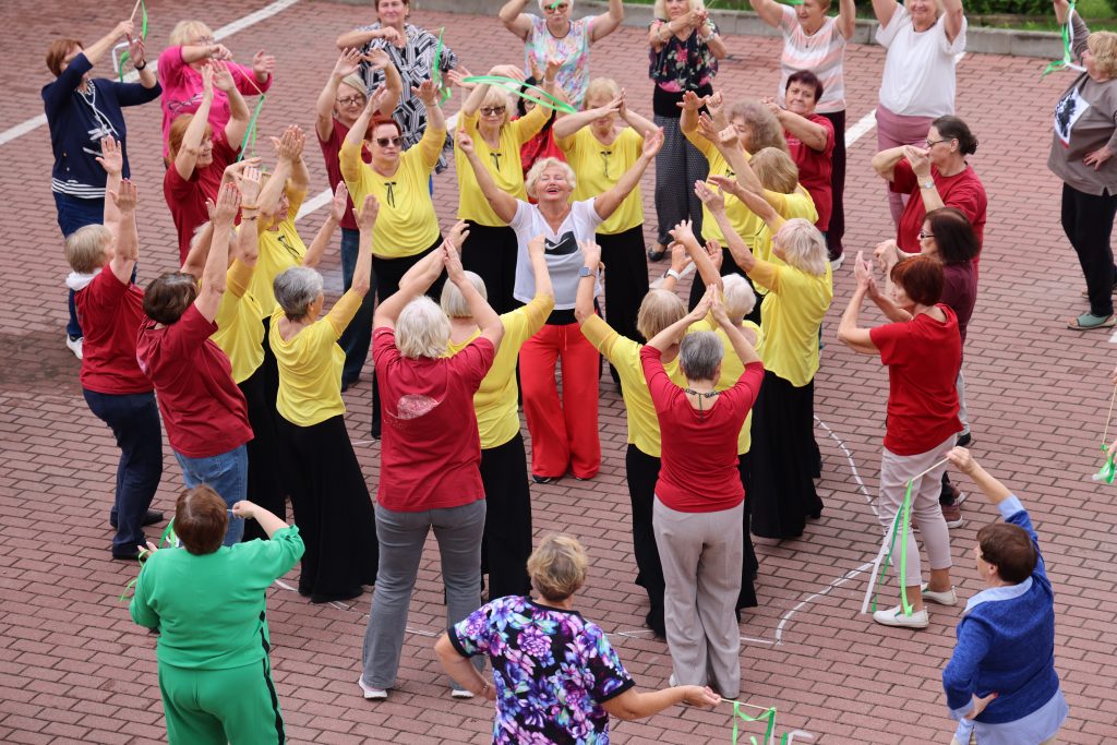 Group of elderly women engaging in a lively dance activity outdoors, wearing colorful shirts, with arms raised in celebration. The event fosters community spirit and promotes physical activity among seniors.