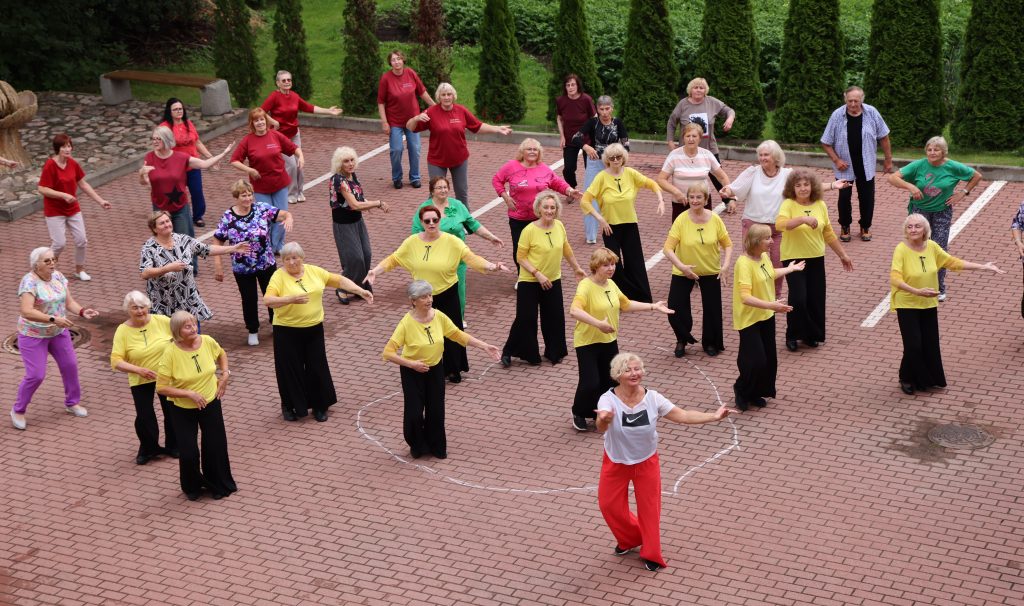 Group of elderly women participating in a lively dance class outdoors, wearing colorful outfits, with some in yellow shirts and others in red, surrounded by greenery and a brick pavement. The scene captures the joy of movement and community engagement in fitness activities.