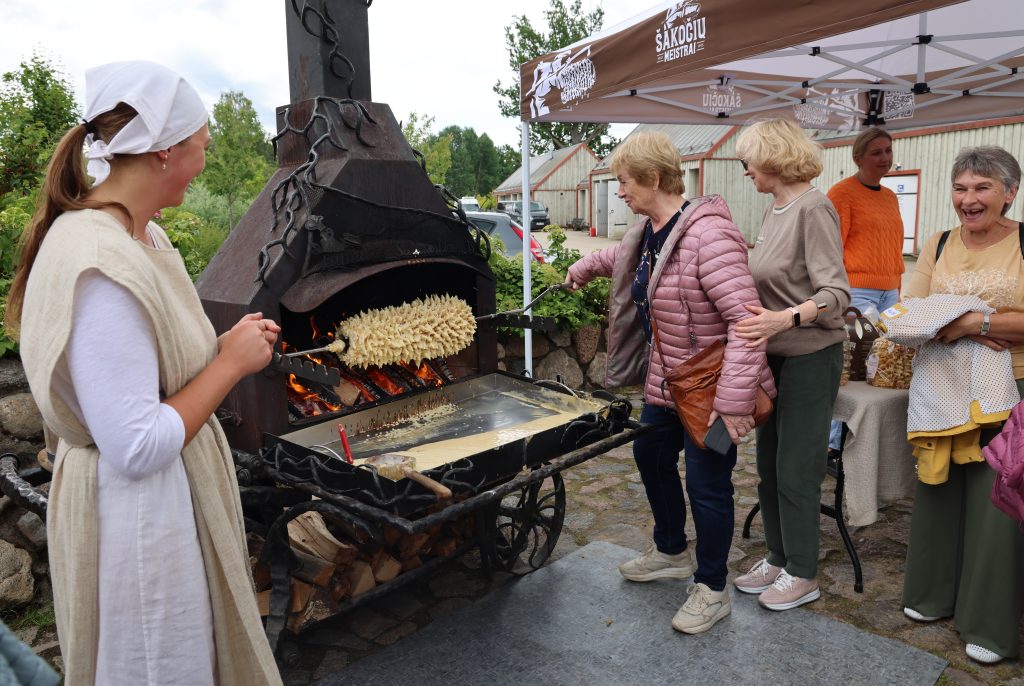 Women participating in a traditional cooking demonstration at an outdoor festival, featuring a large outdoor oven with flames and a wooden skewer of food being prepared. The scene captures the engaging atmosphere of the event, showcasing culinary skills and community interaction.