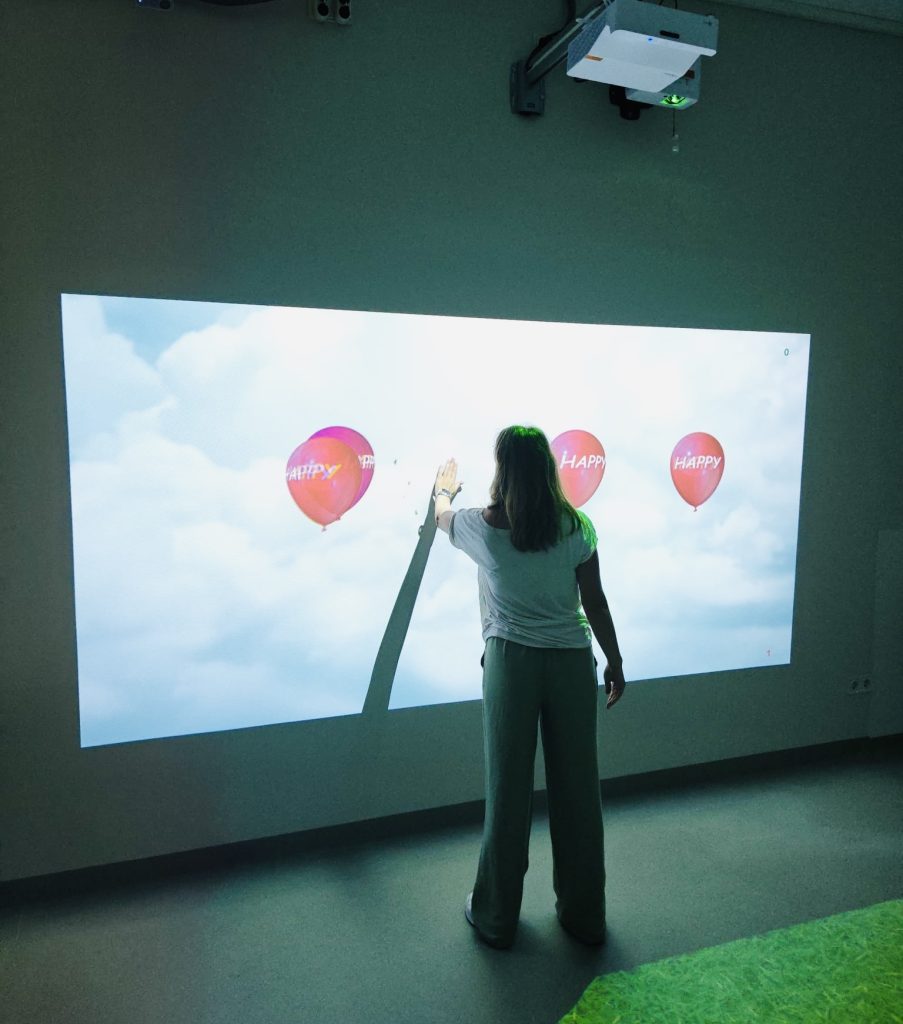 A woman interacts with a projected digital display featuring colorful balloons labeled "Happy," showcasing innovative technology in rehabilitation services as part of the SPEED UP Project.